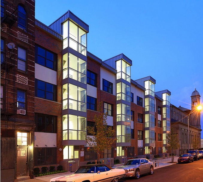 Modern multi-unit residential building at dusk with glowing windows and glass bay towers beside an older brick tenement